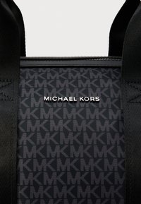 Μαύρη τσάντα ώμου με επαναλαμβανόμενο μοτίβο «MK» σε υφασμένο ύφασμα. Το ασημί λογότυπο «MICHAEL KORS» εμφανίζεται έντονα.