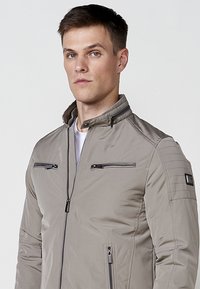 Chaqueta gris claro con un pequeño cuello. Presenta bolsillos con cremallera y detalles cosidos en los hombros. Hecha de un material suave y ligero.