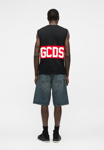 Άνδρας με μαύρο αμάνικο μπλουζάκι με έντονα λευκά γράμματα "GCDS" σε κόκκινη ταινία, τζιν σορτς, μαύρες κάλτσες και μαύρα αθλητικά παπούτσια, γυρισμένος με την πλάτη.