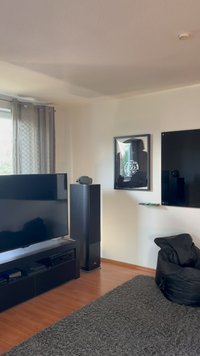 Schwarzer Flachbildfernseher auf einem niedrigen Regal, neben einem hohen schwarzen Lautsprecher. Grauer, plüschiger Teppich, schwarzer Sitzsack, gerahmtes Kunstwerk an der Wand.