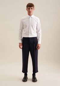 Weißes, langärmeliges Hemd, geknöpft, mit Kragen, kombiniert mit dunkelblauen, plissierten Hosen. Schwarze Schuhe runden das Outfit ab.