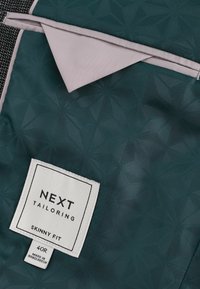 Tela en tono verde azulado con un acabado suave. Un forro de bolsillo beige asoma. La etiqueta dice "NEXT Tailoring, Skinny Fit, 40R, Hecho en Bangladés."