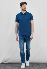 Marinblå polo med tre knappar, kombinerad med ljusblå jeans och vita sneakers, uppvisad på en manlig modell.