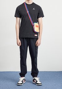 Svart randig t-shirt med en logotyp, lösa svarta cargo-byxor och vita sneakers med röda accenter. Slingväska i gult med blåa/röda remmar.