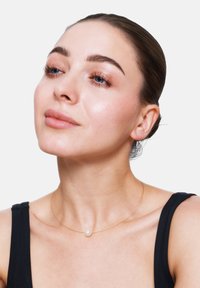 Gouden ketting met een enkele witte parel pendant, met een minimalistisch ontwerp. Model draagt een zwarte mouwloze top met een strakke haarstijl.