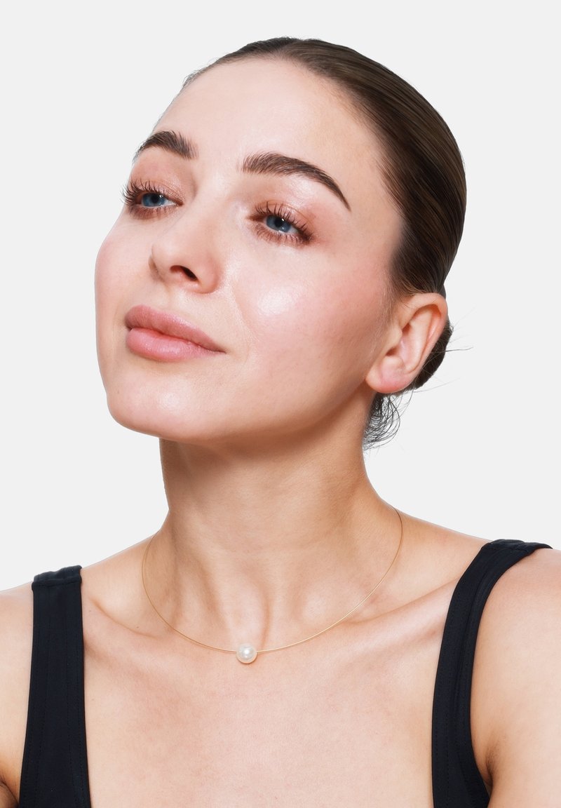 Gouden ketting met een enkele witte parel pendant, met een minimalistisch ontwerp. Model draagt een zwarte mouwloze top met een strakke haarstijl.