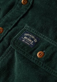 Groen corduroy overhemd met bruine knopen, voorzien van een stoffen patch met het label "SUPERDRY CO MACHINED." Gestructureerd oppervlak, zichtbare stikdetails.