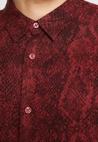 Gros plan d'une chemise rouge à motifs avec un col pointu et trois boutons rouges visibles fermés sur le devant.