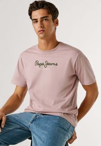 Svetlo roza bombažna majica z zelenim logotipom "Pepe Jeans London" natisnjenim na prsih. Mehka tekstura in standardni kroj.