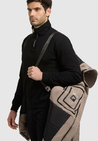 Mustat vetoketjuneuleet, jotka on yhdistetty beigeen ja mustaan eristettyyn takkiin, joka on heitetty olalle. Takissa on tummia yksityiskohtia ja näkyvä etiketti.