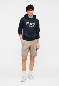 Sötétkék kapucnis pulóver fehér logóval, bézs rövidnadrággal, amely finom, ismétlődő mintával rendelkezik. Fehér zokni és bézs cipő.