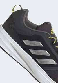 Zapatilla deportiva negra con parte superior texturizada, acentos plateados y una entresuela blanca, con detalles amarillos en el talón y los cordones.