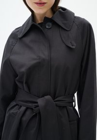 Svart trenchcoat med klassisk krage, framtida knäppning, bälte i midjan och sidofickor. Slät textur med skräddarsydd passform.
