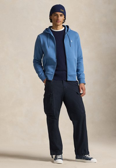 Μπλε φούτερ με φερμουάρ, μπλε πουλόβερ, σκούρο παντελόνι τύπου cargo και αθλητικά παπούτσια. Διαθέτει κουκούλα, μπροστινές τσέπες και ριμπ μανσέτες. Περιλαμβάνεται πλεκτό σκουφάκι.