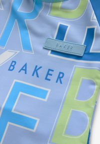 Svetlo modra tkanina za kopalke z velikim besedilom v kontrastni zeleni in modri barvi. Pravokotni označeni trak z napisom "BAKER by Ted Baker."