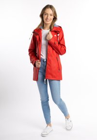 Röd vattentät jacka med huva, elastiska muddar, dragkedja fram och fickor. Kombinerad med ljusblå jeans och vita sneakers.