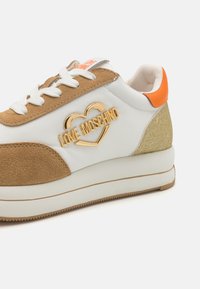 White and tan sneaker with gold "LOVE MOSCHINO" logo, orange accent, textured suede, rubber sole, and lace-up closure.