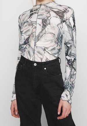 Femme portant un haut transparent à manches longues avec des motifs abstraits noirs, blancs et roses, rentré dans un jean noir taille haute.