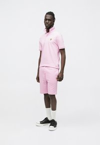 Rausvos spalvos polo marškinėliai ir šortų komplektas, pagamintas iš medvilnės, su mažyčiu geltonu logotipu. Derinamas su juodais sportbačiais ir baltais kojinėmis.