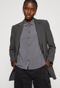 Grijze op maat gemaakte blazer met gestructureerde schouders over een grijze button-up shirt. De outfit is gecombineerd met donkere, gestructureerde broeken.