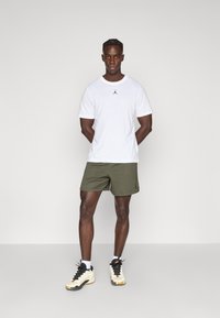 Λευκό t-shirt με λογότυπο Jordan, σορτς σε ελιά πράσινο χρώμα και αθλητικά παπούτσια σε μαύρο και κρεμ χρώμα· μοντέλο στέκεται με τα χέρια πίσω από την πλάτη σε ουδέτερο φόντο.