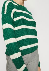 Maglione a righe verdi e bianche in maglia con scollatura tonda, caratterizzato da un tessuto testurizzato e una vestibilità morbida, abbinato a pantaloni grigi.