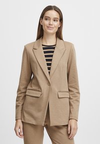 Blazer beige con cierre de un solo botón, dos bolsillos frontales, cuello de solapa y tejido texturizado, llevado sobre una blusa con rayas en blanco y negro.