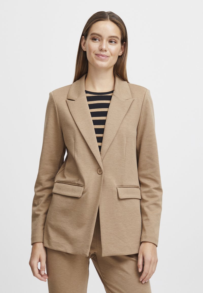 Blazer beige con cierre de un solo botón, dos bolsillos frontales, cuello de solapa y tejido texturizado, llevado sobre una blusa con rayas en blanco y negro.