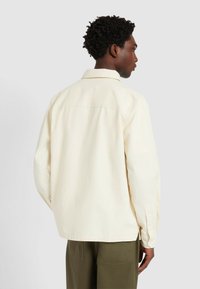 Mężczyzna z krótkimi kręconymi włosami, ubrany w kremową koszulę z długim rękawem i oliwkowe spodnie, stojący tyłem na jednolitym tle.