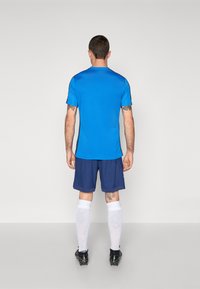 Zila sporta krekls ar īsām piedurknēm, ap kombinēts ar tumši ziliem šortiem un baltām ceļgala garuma zeķēm, vilkti ar melnām futbola kurpēm. Redzami tetovējumi uz rokām.