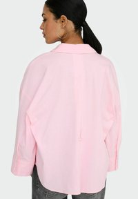 Femme aux cheveux foncés attachés en une queue de cheval basse, portant de grandes boucles d'oreilles en anneau et une chemise ample rose clair à boutons, vue de dos.