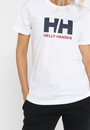 Personne portant un t-shirt blanc Helly Hansen avec un grand logo HH noir et un texte rouge, associé à un pantalon noir.