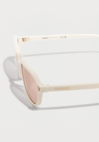 Γυαλιά ηλίου Versace σε κρεμ απόχρωση με φακούς ροζ χρώματος και χρυσές λεπτομέρειες με το λογότυπο στο πλαινό μέρος και τη άρθρωση, σε λευκό φόντο.
