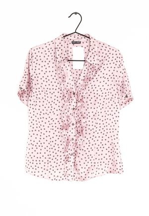 Blouse rose clair à manches courtes avec pois foncés, patte de boutonnage plissée à l'avant et col, suspendue sur un cintre noir.