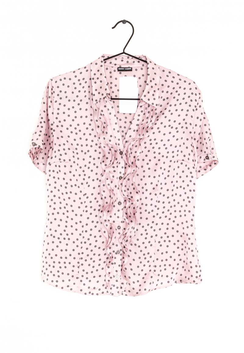 Blouse rose clair à manches courtes avec pois foncés, patte de boutonnage plissée à l'avant et col, suspendue sur un cintre noir.