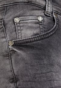 Jeans de mezclilla gris oscuro con remaches metálicos, costuras cosidas y un bolsillo frontal. La textura parece suave con un ligero desgaste.