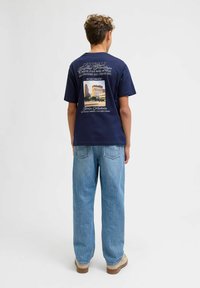 Tamsiai mėlyna medvilninė marškinėliai su grafiniu spausdiniu ant nugaros, vaizduojančiu pastatą, derinami su šviesiai mėlynomis laisvomis džinsomis ir bežiniu avalyne.