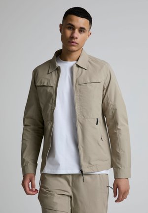 Ung mand iført en beige jakke med lynlås over en hvid t-shirt og matchende beige bukser, stående mod en ensfarvet grå baggrund.