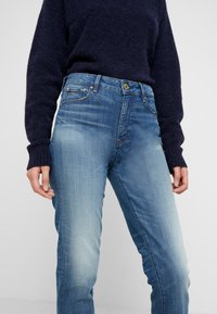 Ljusblå denimjeans med en urtvättad finish, femficksdesign och gula knappdetaljer, ihop med en mörk stickad tröja.
