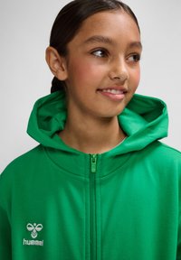 Grøn zip-op hoodie lavet af blødt stof, med en hætte og et lille hvidt logo på venstre bryst. Glat tekstur, enkelt design.