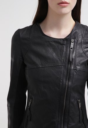 Mujer con chaqueta de cuero negra con cremallera frontal asimétrica y bolsillos laterales con cremallera, sobre un fondo claro y liso.
