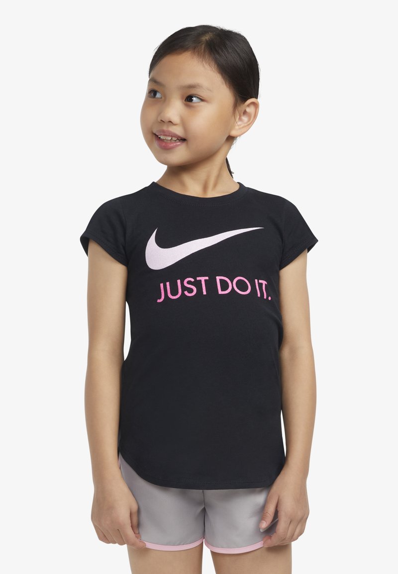 Černé bavlněné tričko s krátkým rukávem, které má velký bílý symbol Nike a růžový text "JUST DO IT." Nosené s šedými šortkami a růžovými akcenty.