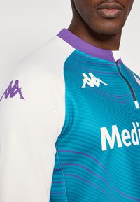 Kappa sportinis marškinėlis, turintis turkio spalvos pagrindą su violetiniais akcentais, aukštą apykaklę ir baltą logotipą. Priekyje yra reljefinis banguotas raštas.