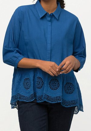 Kobieta w niebieskiej bluzce z długim rękawem z koronkowym wykończeniem na dole i ciemnych spodniach, z delikatnie złożonymi rękami z przodu.