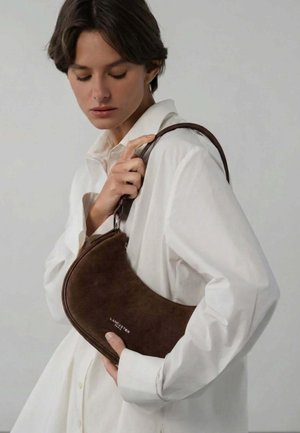 Femme en chemise blanche tenant un sac à main marron en forme de croissant avec bandoulière, marqué "Lancaster Paris."