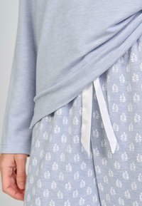 Pantalones de pijama azul claro con patrones de hojas blancas y una cinta blanca, parcialmente cubiertos por una camiseta de manga larga azul claro a juego.