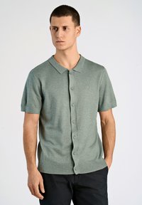 Grön kortärmad polo med krage, knappframsidan och ribbad nederkant. Tillverkad av mjukt, texturerat tyg; med subtil logotypdetalj.