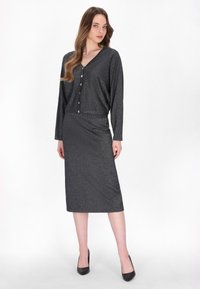 Dunkelgraues, langärmliges Kleid mit V-Ausschnitt und Knopfverschluss, aus einem strukturierten, glitzernden Stoff, reicht bis zur Mitte der Waden und wird mit schwarzen Absätzen kombiniert.