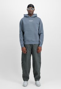 Felpa grigia con testo "STUDIO EDITION", tasca frontale, abbinata a pantaloni cargo verde scuro e scarpe da ginnastica argentate. Occhiali da sole eleganti completano il look.