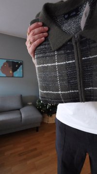 Suéter con cremallera de cuadros verdes y grises, con cuello abotonado, hecho de tela texturizada, sostenido en las manos contra un fondo neutro.